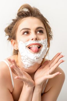 Portrait of a beautiful young smiling caucasian woman posing with shaving foam on the face stuck out her tongue on white background