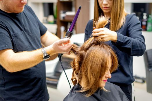 Two hairstylists using curling iron on customers long brown hair in a beauty salon