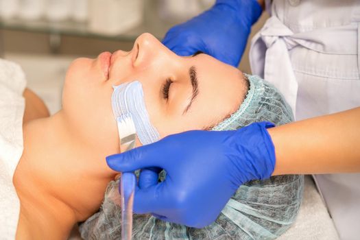 Beautician covering woman facial skin with moisturizing cleansing mask during skin care procedure in a spa beauty salon