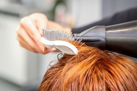 Closeup of master's hand with blow-drying and hairbrush blowing female red hair in a salon