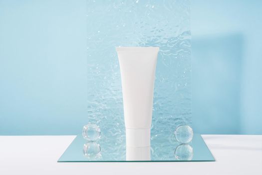 Unbranded cosmetic cream white plastic tube mockup on blue background with stylish props, mirror and glass balls, acrylic plate. Body and health care beauty product packaging. Product presentation