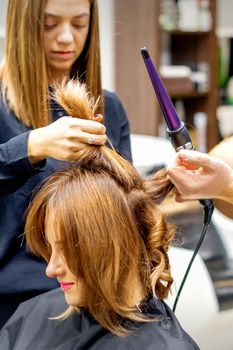 Two hairstylists using curling iron on customers long brown hair in a beauty salon