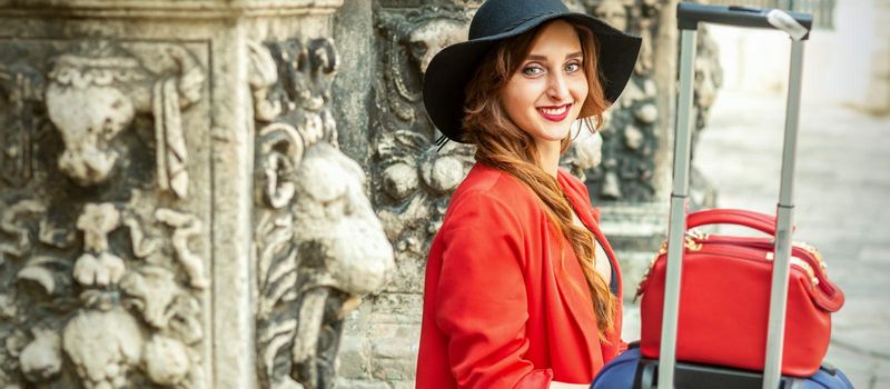 Portrait of a beautiful smiling young caucasian woman in a black hat with luggage sitting at the ancient building looking at camera outdoors