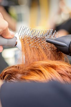 Closeup of master's hand with blow-drying and hairbrush blowing female red hair in a salon