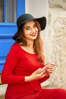 Portrait of tourist young caucasian woman in a red long dress and black hat with suitcase sitting on the stairs outdoors