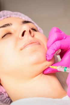 The cosmetologist makes lipolytic injection on the chin of a young woman against the double chin in a beauty salon