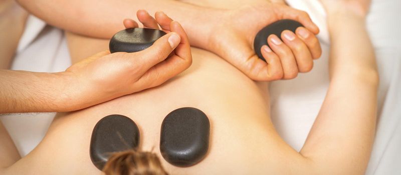 Hot stone massage on the female back with hands of masseur holding black massage stones in spa salon