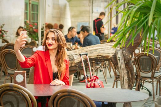 Tourist young caucasian woman in a red jacket with suitcase takes a selfie at the table in cafe outdoors