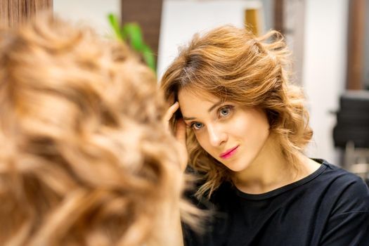 The beautiful young caucasian woman looks at her reflection in the mirror checking hairstyle and makeup in a beauty salon