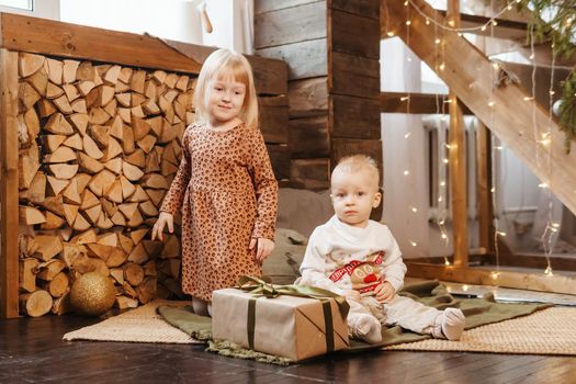 Little brother and sister play on Christmas eve in a beautiful house decorated for the New Year holidays. Children are playing with a Christmas gift. Scandinavian-style interior with live fir trees and a wooden staircase.
