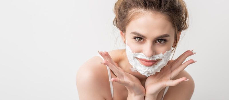 Portrait of a beautiful young smiling caucasian woman with shaving foam on face poses on white background