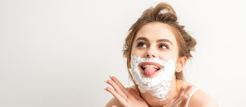 Portrait of a beautiful young smiling caucasian woman posing with shaving foam on the face stuck out her tongue on white background