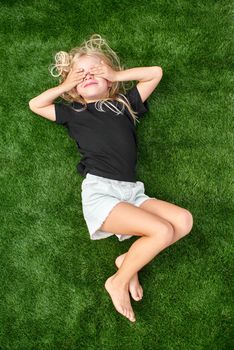 Top view. Mock up. Pretty blonde child girl with closed eyes lying, relaxing, stretching on green grass. Smiling preschool girl 5 - 6 years old in black t shirt. Lifestyle. Summer vacations. Leisure