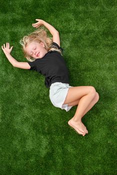 Top view. Mock up. Cute blonde child girl with closed eyes lying, relaxing, stretching on green grass. Smiling preschool girl 5 - 6 years old in black t shirt. Summer vacation. Leisure. People concept