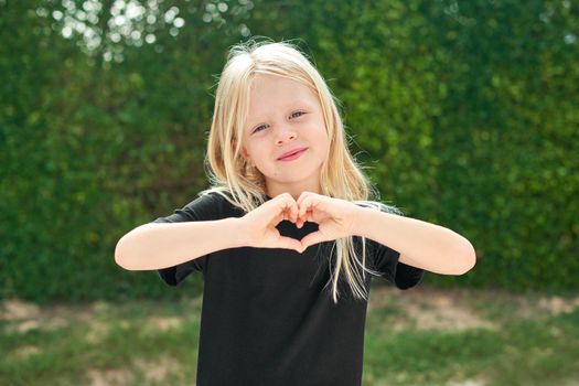 Mock up. Pretty blonde child girl showing sign of love heart from fingers hands before green wall background. Smiling preschool girl 5 - 6 years old in black t shirt looking at camera Summer vacations