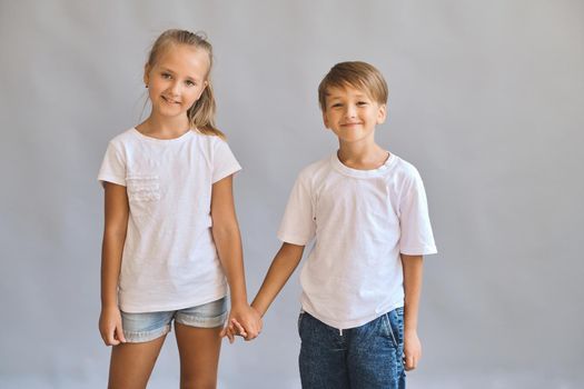 Cute smilling two kids, little boy and girl in white t-shirts hold hands best friends on gray background. Blank white t-shirt for your design