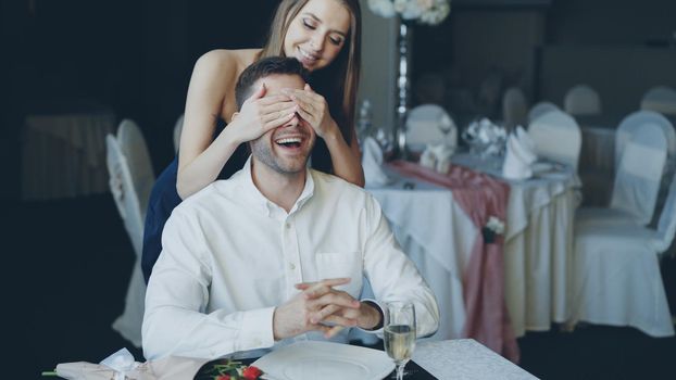 Pretty young woman is greeting her boyfriend in restaurant closing his eyes with her hands during dating