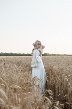 A blonde woman in a long white dress walks in a wheat field. The concept of a wedding and walking in nature.