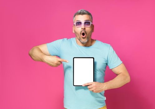 Great offer pointing at digital tablet white screen handsome man looking at camera with shock facial expression wearing casual blue shirt and sunglasses isolated on pink background. App advertisement.