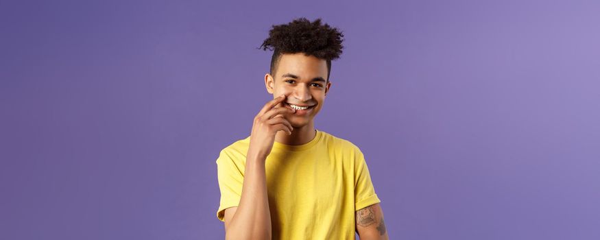 Close-up portrait of lovely young hispanic gay man with dreads, tattoos, touching lip sensually and flirty smiling, checking out someone really cute and handsome, standing purple background.