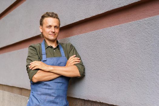 Handsome employee man standing outside with arms folded wearing apron take a break leaned on a wall enjoying fresh air after long hard day. Freelancer man at coffee brake enjoying free time.