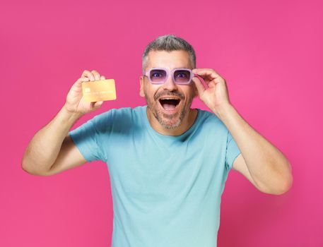 Excited, shocked handsome middle aged man hold debit, credit card in hand fixing his glasses wearing blue t-shirt isolated on pink background. Financial, banking concept.