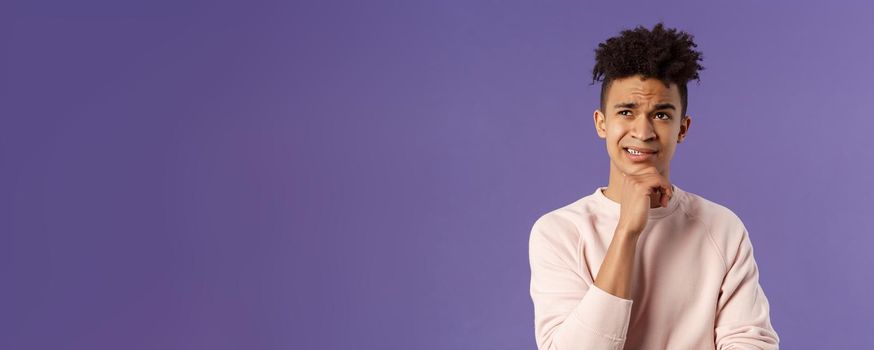 Close-up portrait of indecisive, puzzled young man facing difficult choice, look up thoughtful, touch chin and grimacing while thinking, making-up idea or decision, purple background.