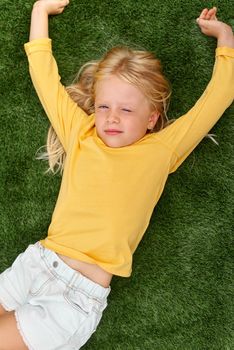 Top view. Mock up for logo, text, design. Blonde long hair child girl lying, stretching on green grass. Preschool girl 5-6 years old in yellow t shirt. Lifestyle Summer vacation Leisure. People