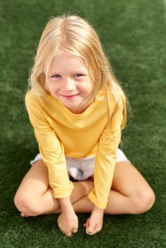 Top view. Pretty beautiful blonde child girl sitting on green grass. Smiling preschool girl 5 - 6 years old in yellow t shirt looking at camera. Lifestyle. Beauty. Summer vacation. leisure. people
