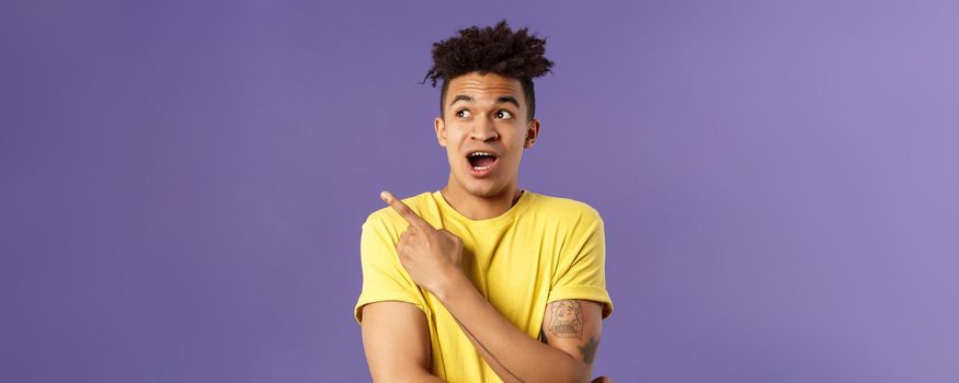 Portrait of impressed hispanic guy seeing something interesting, pointing finger and looking upper left corner, discuss strange thing upwards, standing purple background curious.
