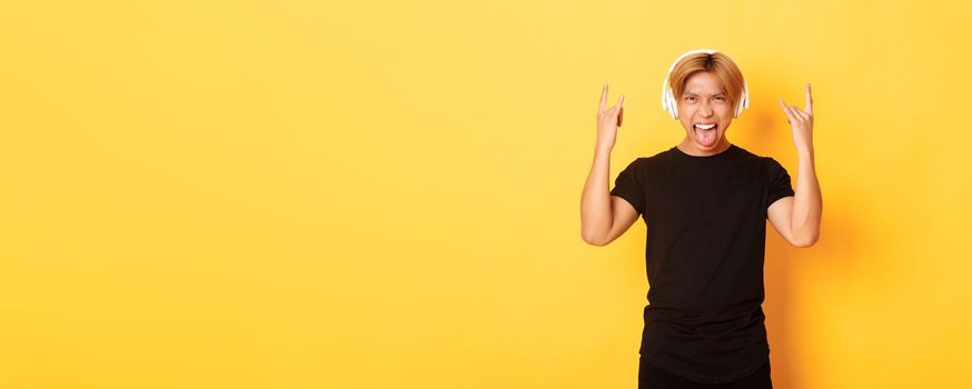 Sassy handsome asian guy likes heavy metal, listening hard rock music, showing rock-n-roll gesture and wearing wireless headphones, sticking tongue, standing yellow background.