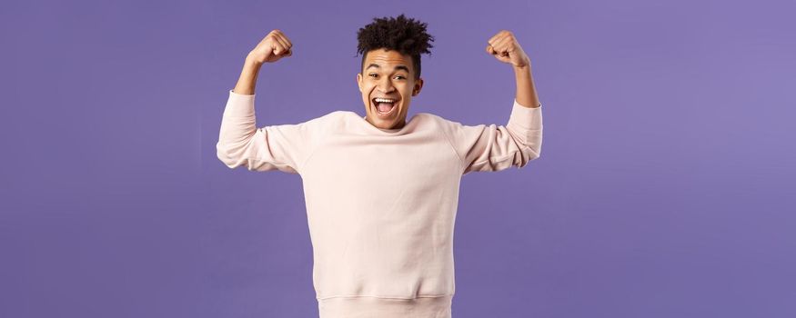 Portrait of young happy man got scholarship, applied to cool university, raise hands up flex biceps like champion, triumphing from great news, achieve goal and rejoicing, purple background.