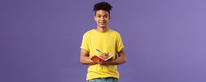Portrait of young hispanic male student studying online courses, writing down lecture, making personal schedule or taking notes in planner, look camera enthusiastic and interested, purple background