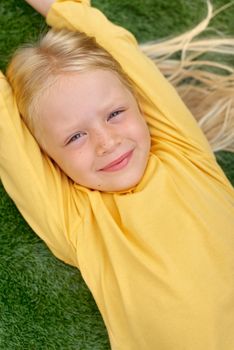 Top view. Mock up for logo, text, design. Blonde long hair child girl lying, stretching on green grass. Preschool girl 5-6 years old in yellow t shirt. Lifestyle Summer vacation Leisure. People