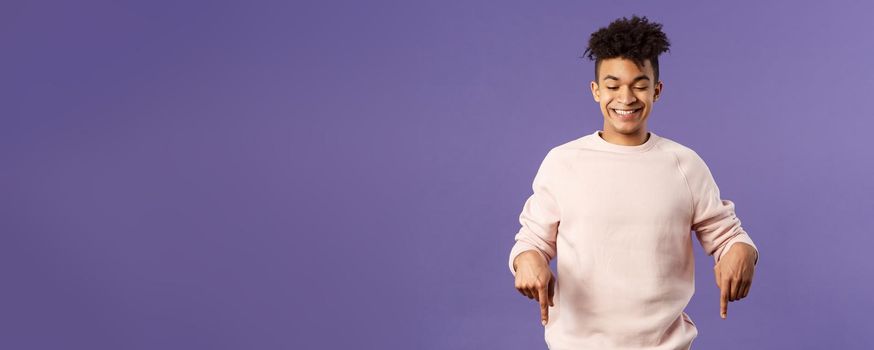 Portrait of cute enthusiastic young man found something really cool, pointing fingers down, happy smiling and peeking at bottom advertisement with pleased grin, purple background.