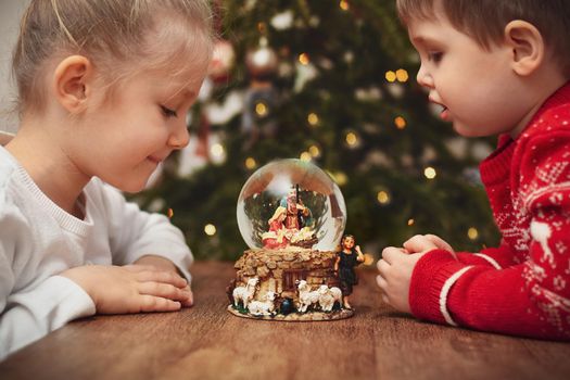 Children looking at a glass ball with a scene of the birth of Jesus Christ near christmas tree