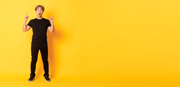 Full-length of impressed asian guy in black clothes, gasping amazed and pointing fingers up, yellow background.