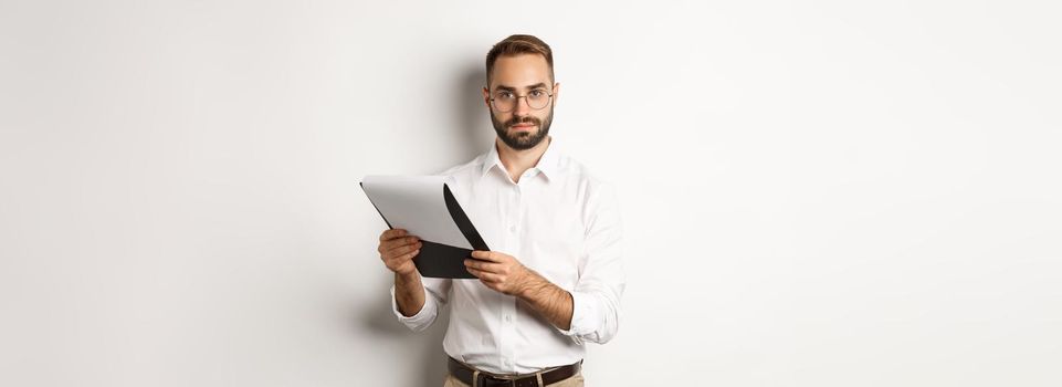 Serious employer looking at camera while reading documents on clipboard, having job interview, standing over white background.