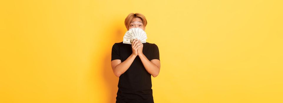 Excited lucky blond asian guy rejoicing from winning cash, holding money and looking happy, standing yellow background.