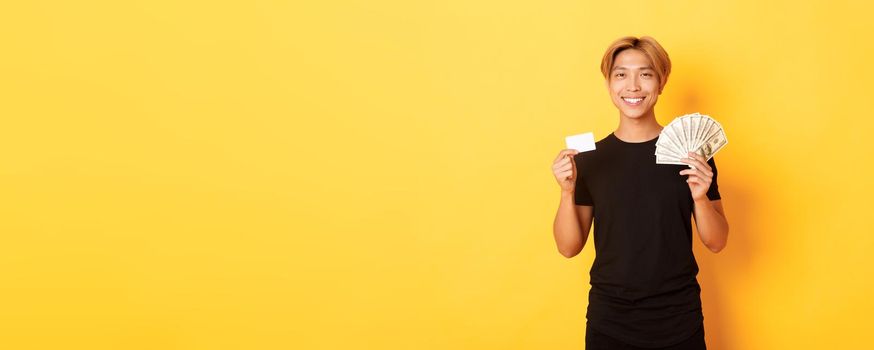 Pleased handsome asian guy showing money and credit card, smiling happy, standing yellow background.