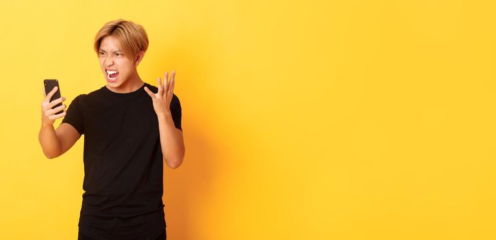 Portrait of angry and pissed-off asian man looking mad at smartphone screen, having argument during video call, standing yellow background.