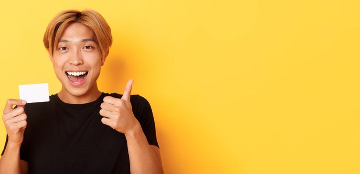 Close-up of happy and satisfied asian handsome guy, showing credit card and thumbs-up in approval, smiling amazed, standing yellow background.