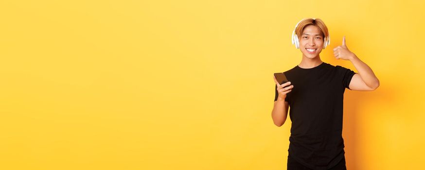 Happy satisfied asian guy likes music or podcast, showing thumbs-up in approval, holding mobile phone, standing yellow background.