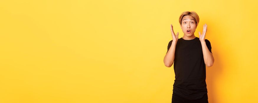 Portrait of surprised gasping blond asian guy reacting to awesome news, yellow background.