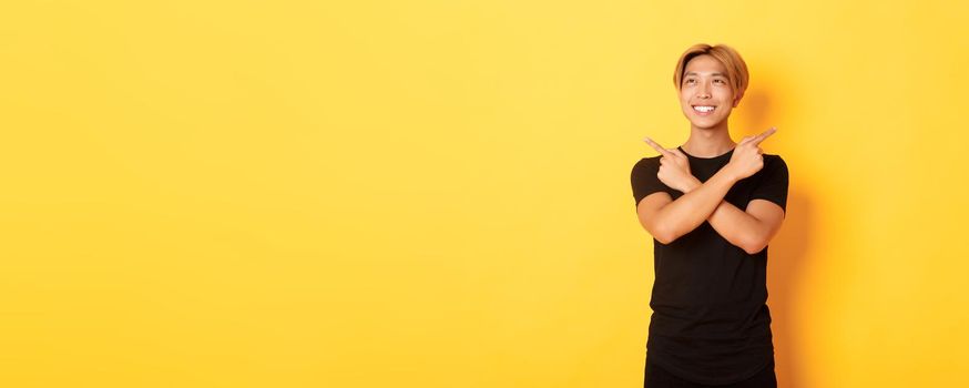 Portrait of smiling pleased asian guy, looking left and making decision, pointing sideways, yellow background.