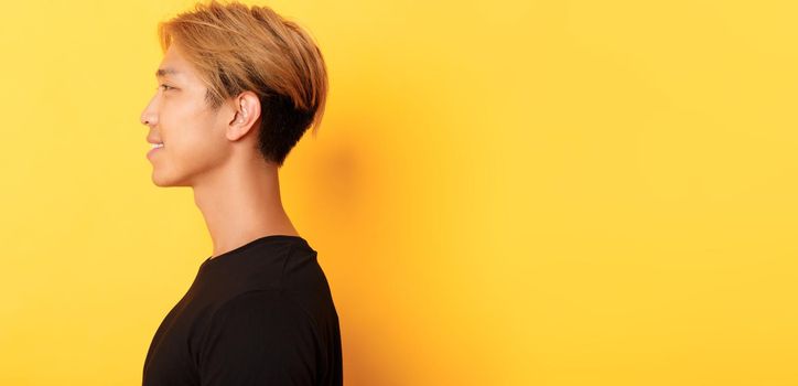Profile of stylish handsome asian guy with fair hair looking left and smiling, standing over yellow background.