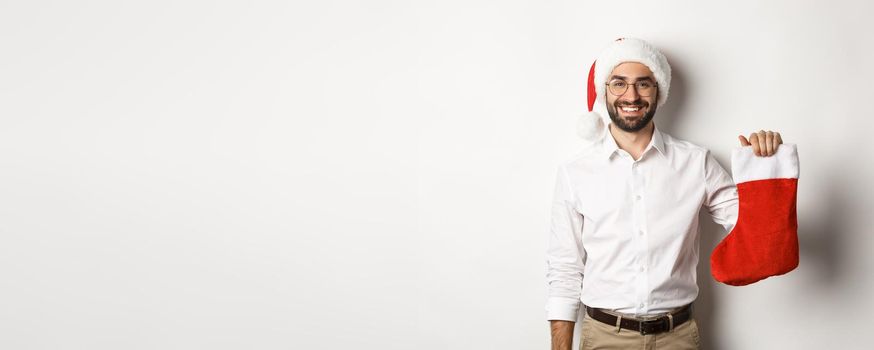 Merry christmas, holidays concept. Happy adult man receive gifts in xmas sock, looking excited, wearing santa hat, white background.