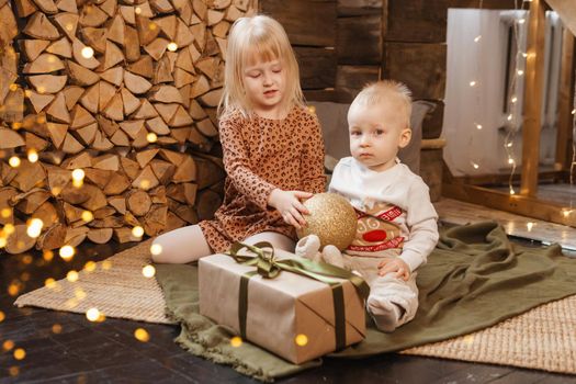 Little brother and sister play on Christmas eve in a beautiful house decorated for the New Year holidays. Children are playing with a Christmas gift. Scandinavian-style interior with live fir trees and a wooden staircase.