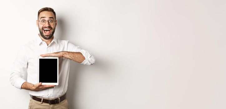 Excited man showing digital tablet screen, smiling amazed, standing over white background.