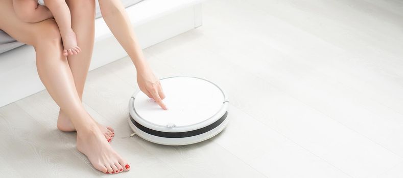 Young mother sitting on sofa with toddler son and watching robot vacuum cleaner doing housework